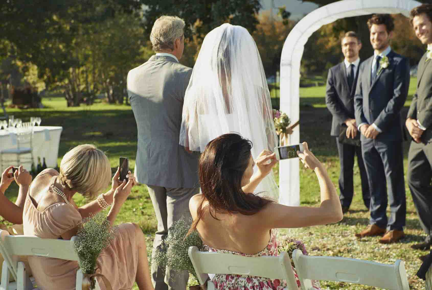 Rear view shot of a father walking his daughter down the aisle while guests take photos with their cellphones